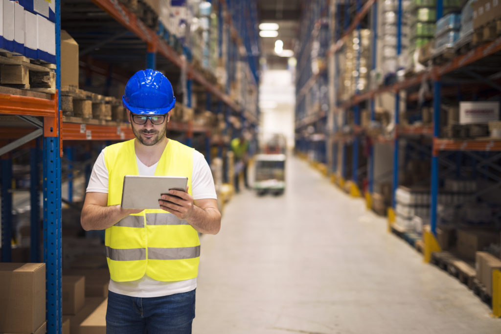 Warehouse worker checking inventory on his tablet while walking in large storage department with shelves and packages in background.